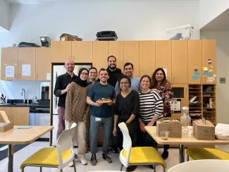 A group of nine young people pose in a kitchen setting holding a birthday cake and smiling