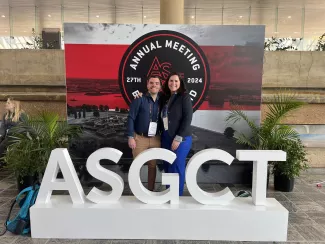 Two people pose against a backdrop advertisement for the ASGCT Annual Meeting