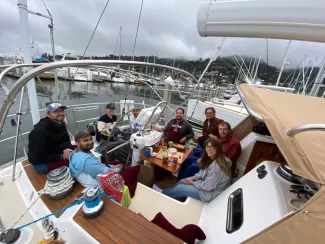 Group of people in the hull of a sailing boat look up at the camera and smile while enjoying chips and drinks with various other sailboats in the background of the marina